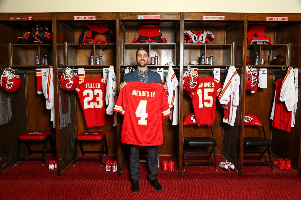 Ben in a blazer holding his personalized red Chiefs jersey, NIEHUES IV number 4, in the Arrowhead locker room between the Fuller and Mahomes stalls.