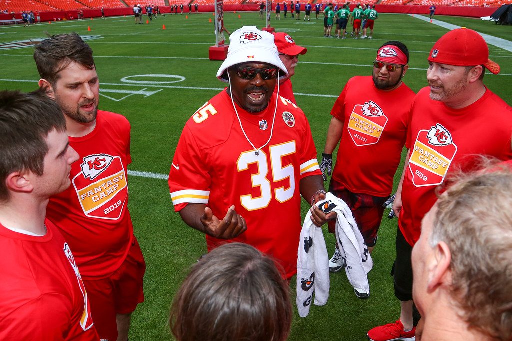 Ben and his team in a huddle on the field as Christian Okoye, in a number 35 jersey and white Chiefs bucket hat, gives a pep talk.
