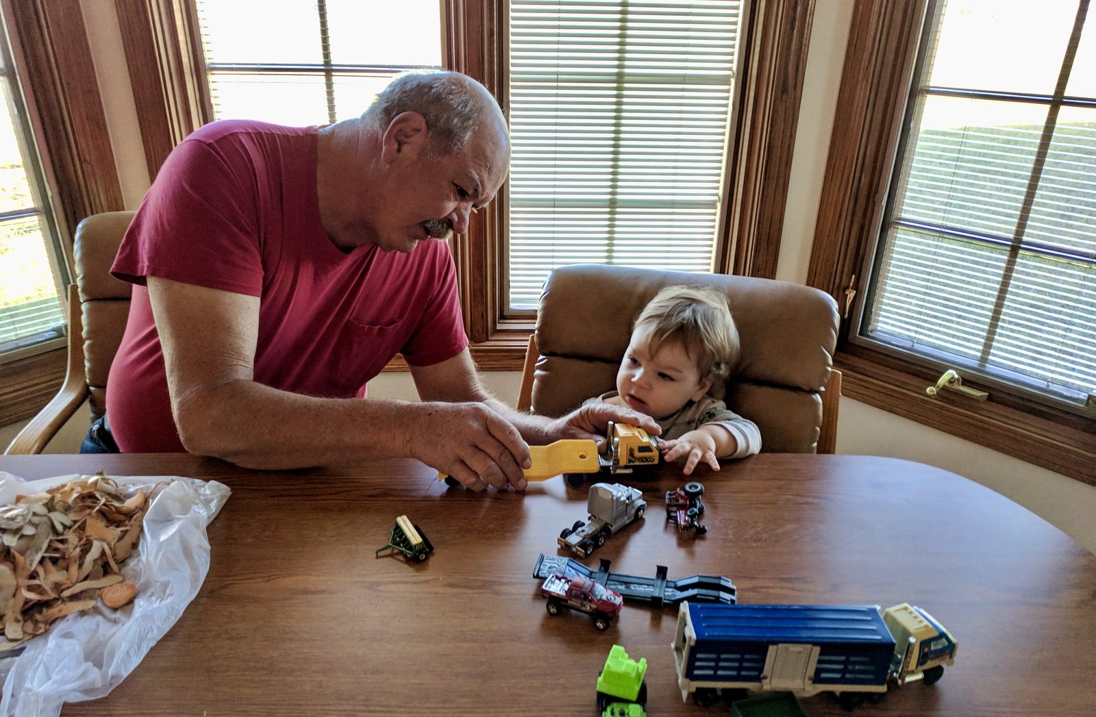 My dad leaning across the Thanksgiving table, handing his young grandson a toy truck. Potato peels in a bag, a spread of toy trucks between them.
